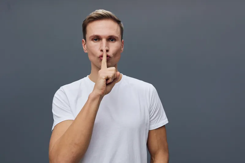 Man with fair skin and short brown hair wears casual white t-shirt, shows hush gesture with index finger over lips, stands against plain gray background, emphasizing silence and secrecy, studio portra
