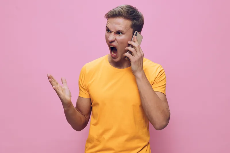 Young man expressing frustration or anger while talking on a mobile phone, wearing a bright yellow t-shirt against a pink background. Casual clothing, emotional reaction, studio shot, vibrant colors. 