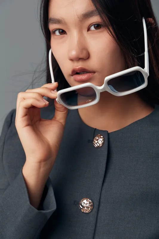 Young woman holding eyeglasses near face with focused expression wearing dark gray garment with decorative buttons studio portrait with neutral background and natural makeup.
