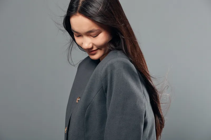 Young woman with long hair wearing gray coat looking down with slight smile on gray background. Casual portrait with natural pose and soft lighting for fashion or lifestyle concept.