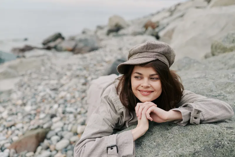 Young woman in knit sweater and hat lies on a large rock on a rocky beach, smiling softly. Overcast sky and muted tones create a calm, peaceful outdoor atmosphere.