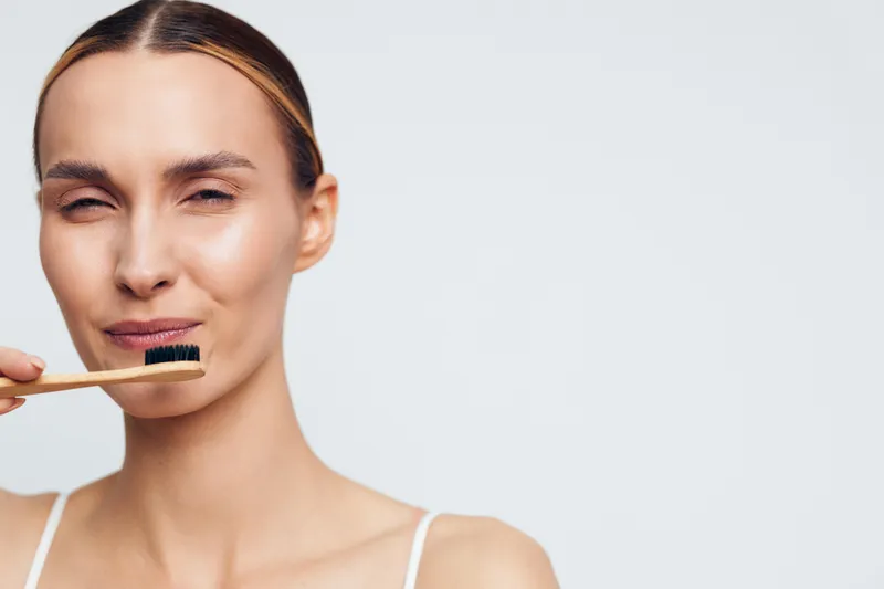 Young woman with clear skin holding natural toothbrush near face, minimal makeup, neutral background, skincare routine, personal care, healthy lifestyle concept.