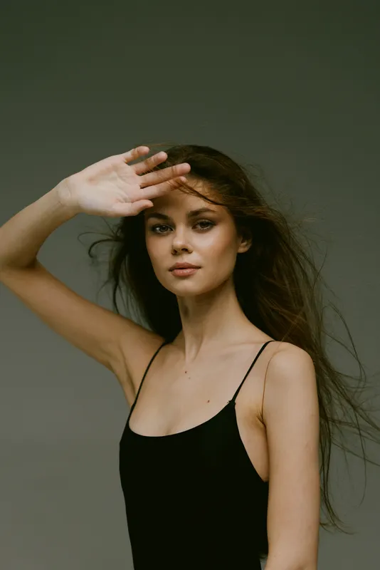 young woman with long hair shielding eyes, wearing black tank top, neutral expression, natural light portrait