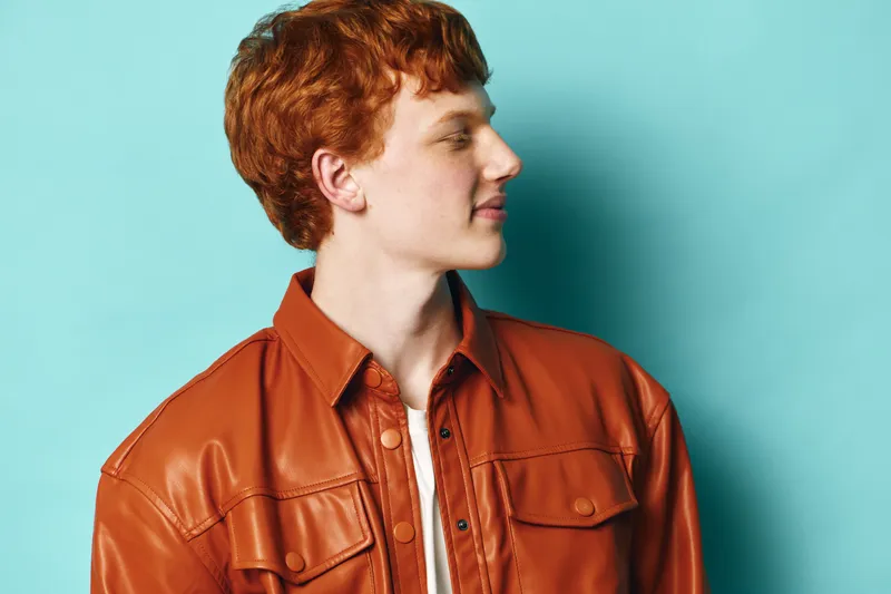 Young man with red hair wearing brown leather jacket and white shirt stands against blue background looking sideways with profile view for casual fashion portrait.