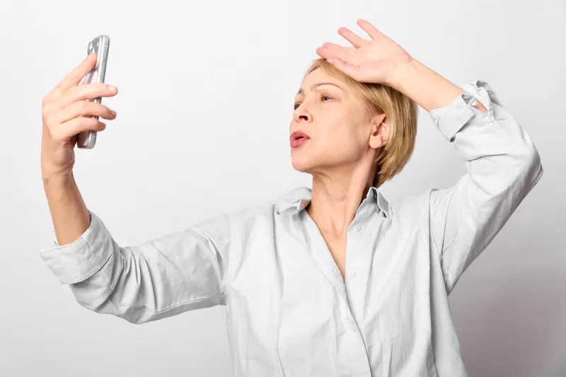 Woman holds smartphone for selfie, covering forehead with one hand, showing headache and stress in studio, white background, portrait of exhausted female, natural light, closeup