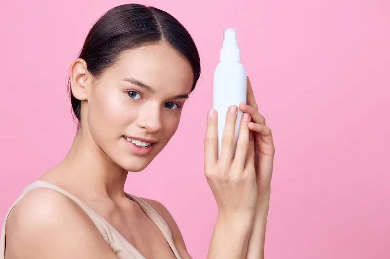 A young woman of Asian descent holding a white skincare bottle against a pink background, smiling with confidence and showcasing her healthy skin for beauty and skincare concepts