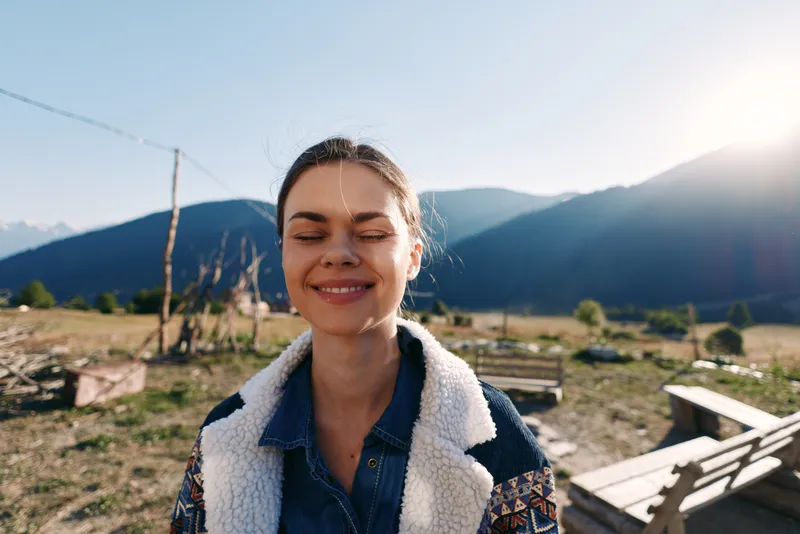 Woman smile mountains outdoors sunlight meadow portrait closed eyes, young female enjoying nature with peaceful expression and warm backlight, casual denim jacket in sunlit alpine field.