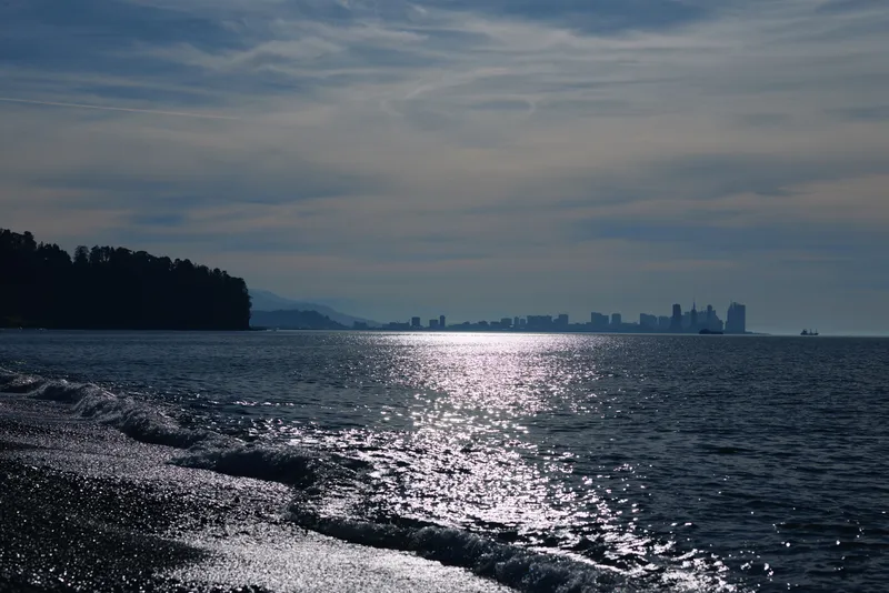 Calm sea reflects bright sunlight toward a distant skyline, with a dark shoreline on the left and gentle waves across the water, creating a tranquil coastal scene at dusk.