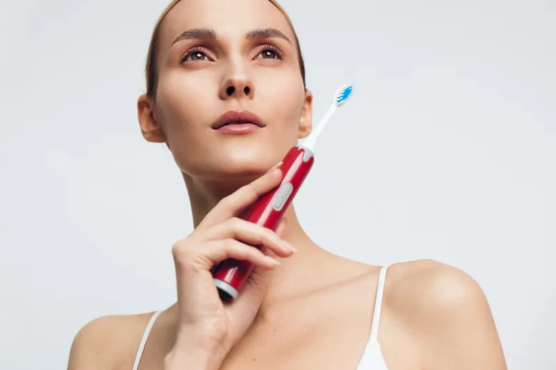 Young woman with clear skin holding electric toothbrush near face on white background. Concept of dental hygiene, skincare, and morning routine. Soft lighting highlights natural beauty.