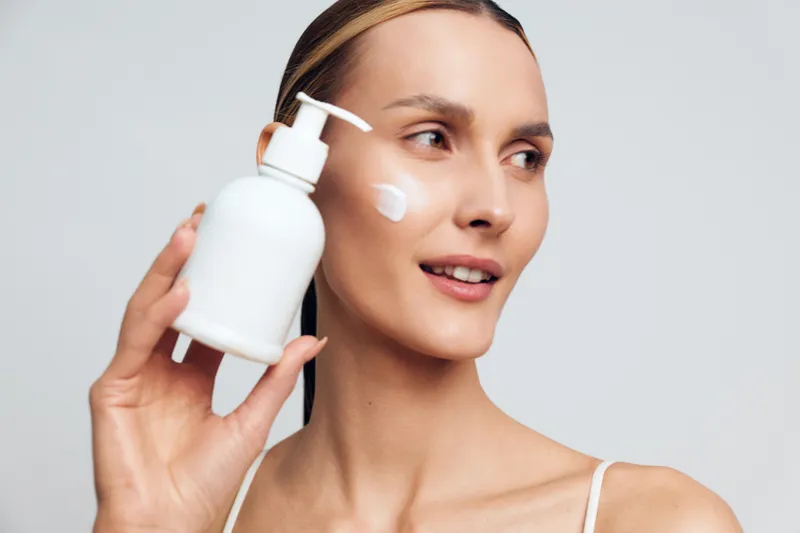A young woman with brunette hair applies lotion from a white pump bottle in a close-up portrait, conveying a skincare routine and self-care in a clean studio.