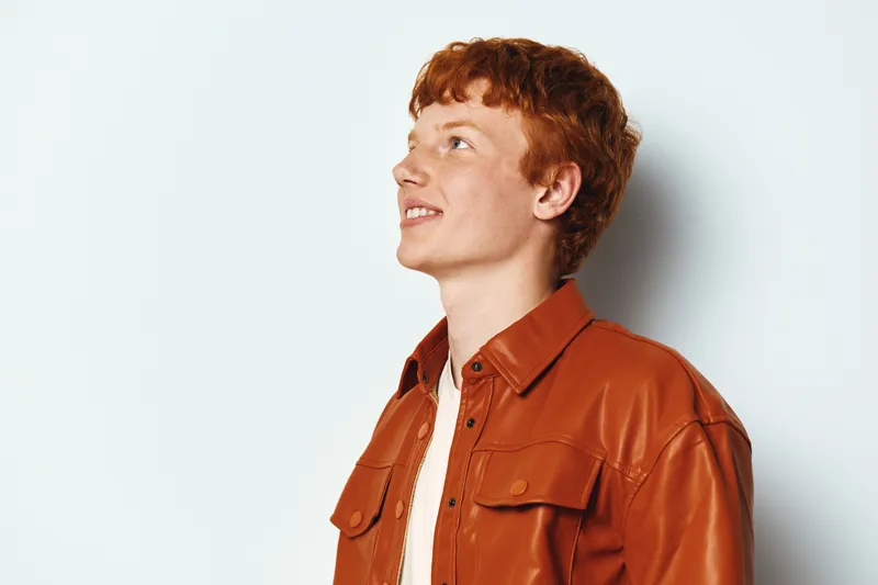 Young man with red hair wearing orange leather jacket looking sideways with slight smile on white background, casual style and confident expression.