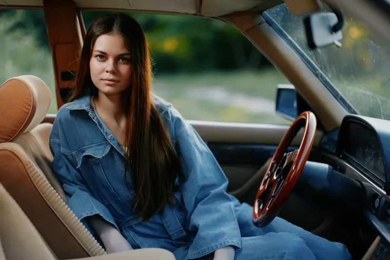 Young woman in denim outfit sitting in vintage car, showcasing a calm and confident expression in a natural outdoor setting, perfect for lifestyle and automotive themes.