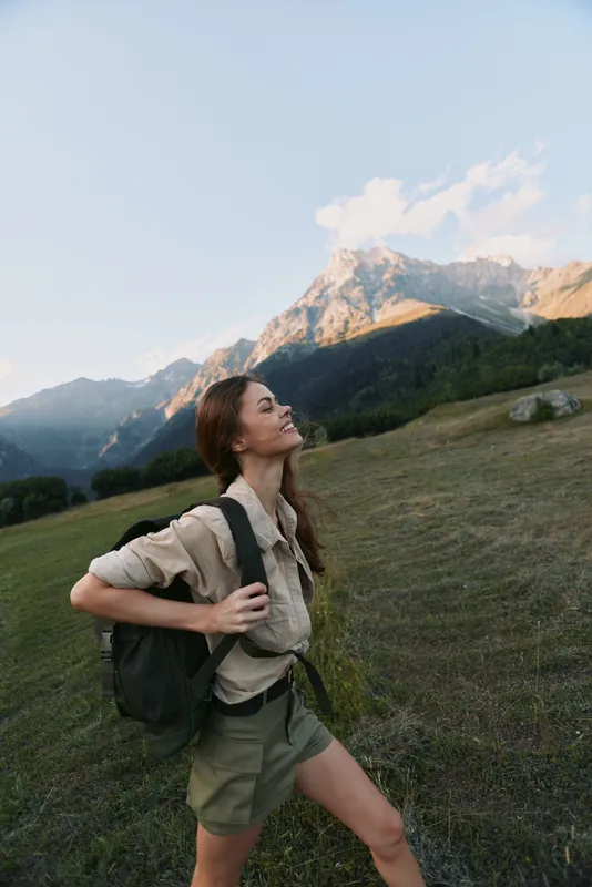 A woman hiker in casual gear stands on a grassy hillside, backpack secured, gazing toward distant mountains under a clear blue sky. A lifestyle moment of outdoor exploration.