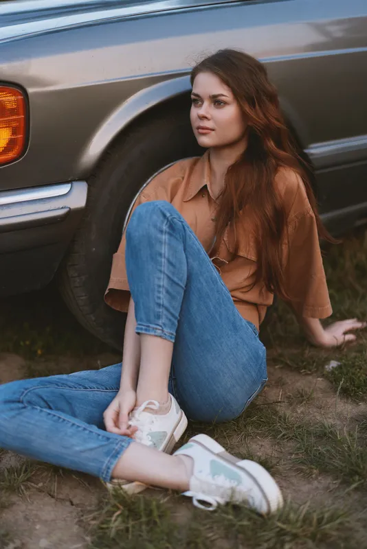 Young woman sitting by a vintage car, wearing casual clothing, expressing a thoughtful mood, with a warm color palette and a natural background.