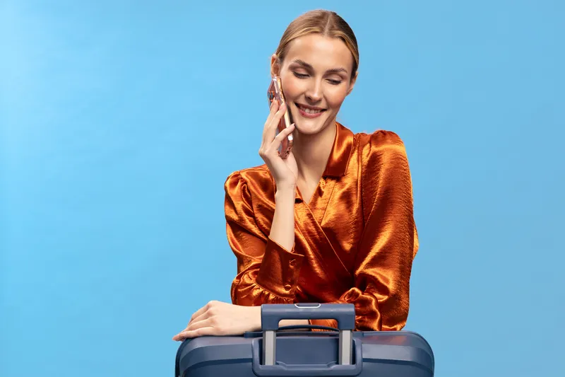 Blonde woman in shiny orange blouse smiles gently with eyes closed, touching face while leaning on blue suitcase, isolated on light blue background. Concept of travel, relaxation, and happiness.