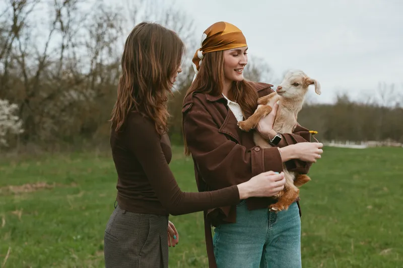 Two women outdoors interact with a small goat, smiling as they cradle the playful kid, capturing a candid countryside lifestyle moment with friendly connection and natural setting.