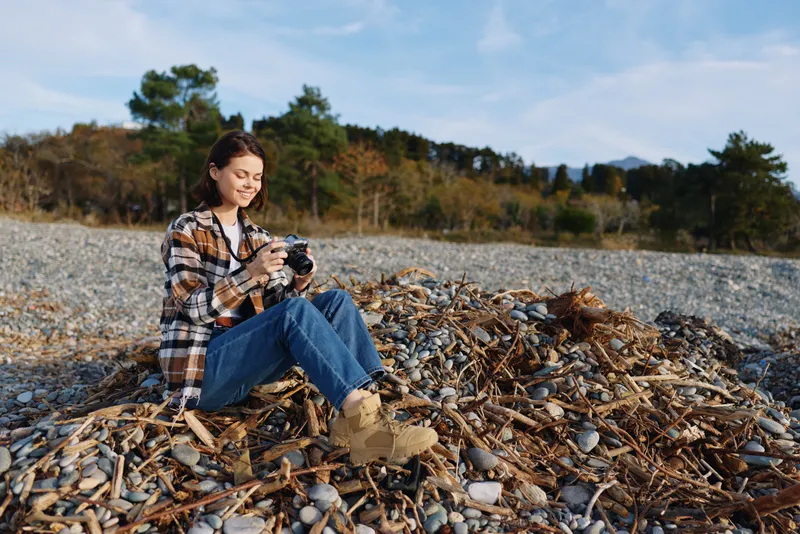 Outdoor woman with a camera sits on a rocky beach among driftwood wearing a plaid shirt and jeans focused on capturing natural scenery and coastal landscape details