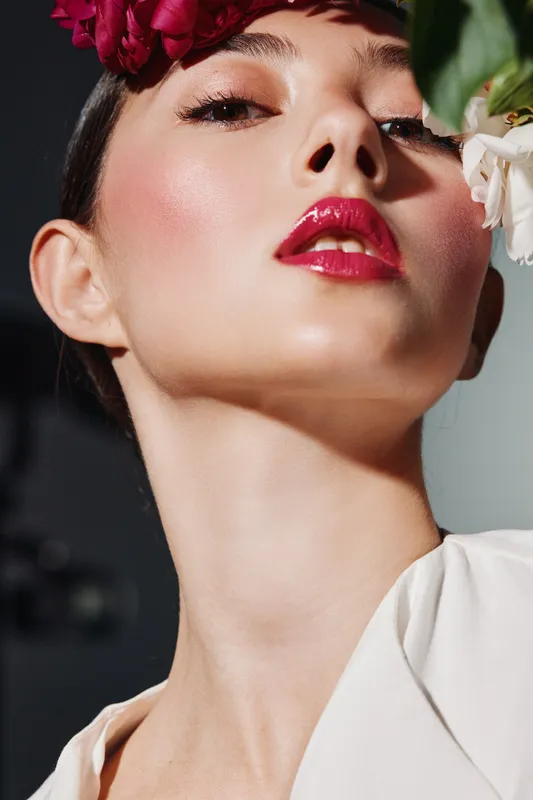 Close-up portrait of a fashion model with glossy red lips and floral accessories. Her radiant skin and sharp gaze create a bold beauty concept and modern elegance.
