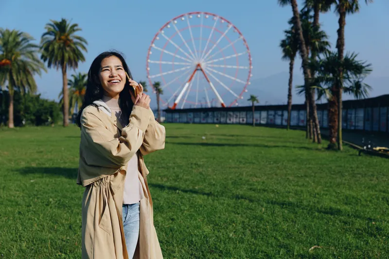 Smiling woman in a light beige coat stands on a sunny park lawn with a ferris wheel in the distance, capturing a candid moment of leisure, outdoor exploration and carefree urban activity