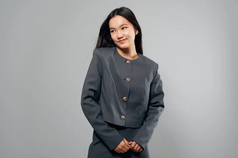 Young woman posing and smiling in formal grey jacket with buttons against plain background for fashion, portrait, business and confidence concept indoors.