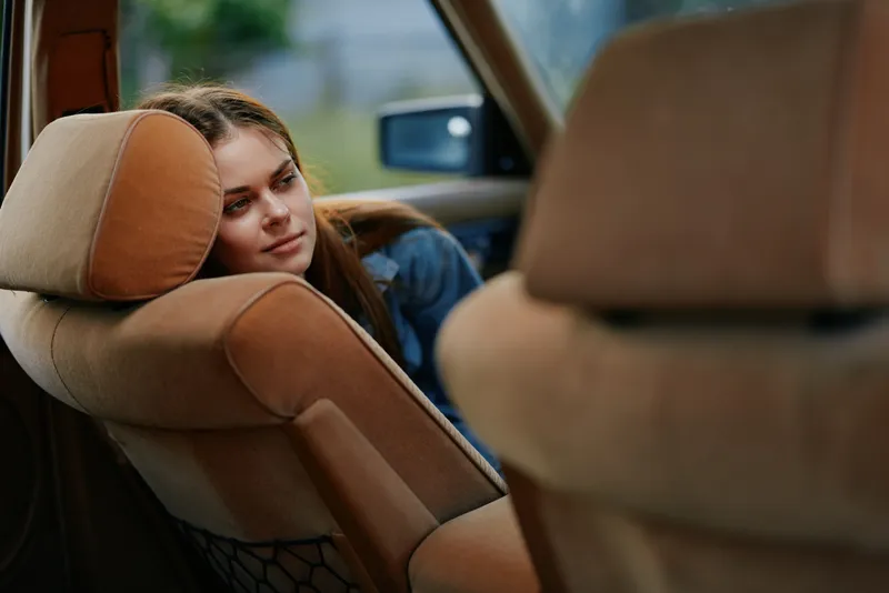 Young woman with long hair resting in a car seat, feeling relaxed and thoughtful, with soft natural light and earthy color tones, ideal for lifestyle and travel themes.