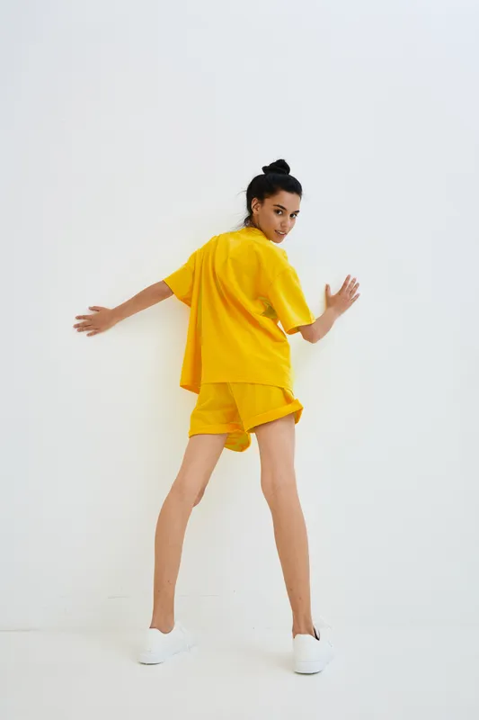 Young woman wearing bright yellow casual outfit poses against white wall, showing playful, sporty style with white sneakers in minimalistic studio setting.