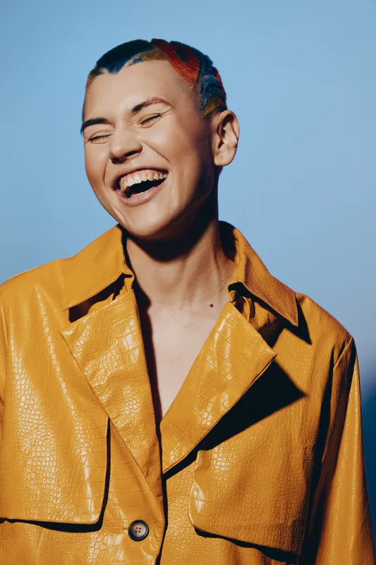 laughing woman with colorful short hair wearing a vibrant yellow jacket, expressing joy and energy against a simple blue background, youthful and lively mood