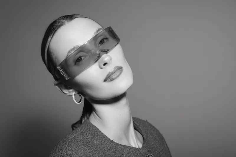 Portrait of a white woman with pale skin wearing transparent sunglasses and hoop earrings. Studio shot in black and white with high contrast lighting and neutral background.