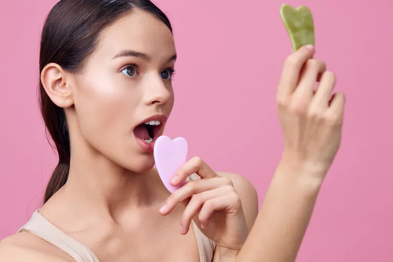 Young woman using beauty tools, smiling and enjoying self care with gua sha stones against a pink backdrop, emphasizing wellness and skincare routines