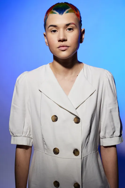Portrait of a young woman with a colorful buzz cut wearing a white buttoned dress, standing against a vibrant blue background, displaying a calm and confident expression.