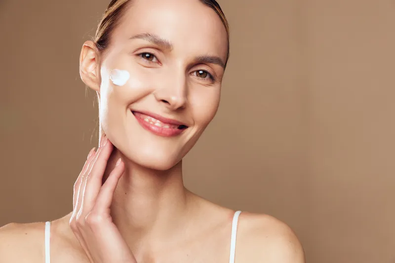 Smiling woman applies moisturizing cream on face with hand isolated on beige background natural skin care beauty routine concept closeup portrait of happy female with clean healthy complexion.
