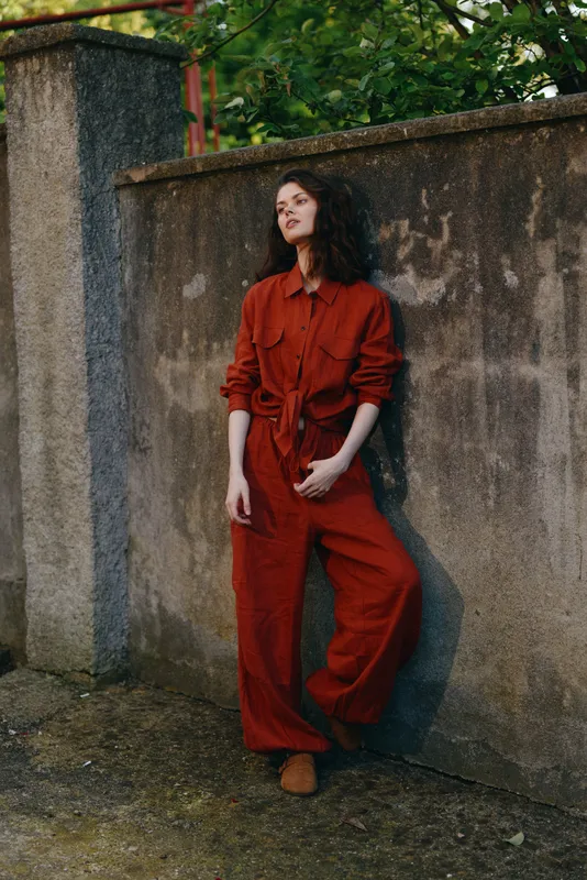 Woman in red jumpsuit leans against a weathered wall in an outdoor urban setting, radiating casual confidence, relaxed mood, and contemporary lifestyle fashion for a polished