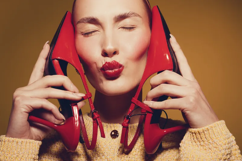 Young woman with closed eyes holds red high heels near face, wearing yellow sweater. Concept of fashion, style, beauty, and footwear in studio with warm lighting and playful mood.