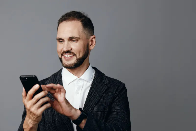 A professional man in business attire engages with a smartphone, displaying confident and positive emotions against an isolated colorful background.