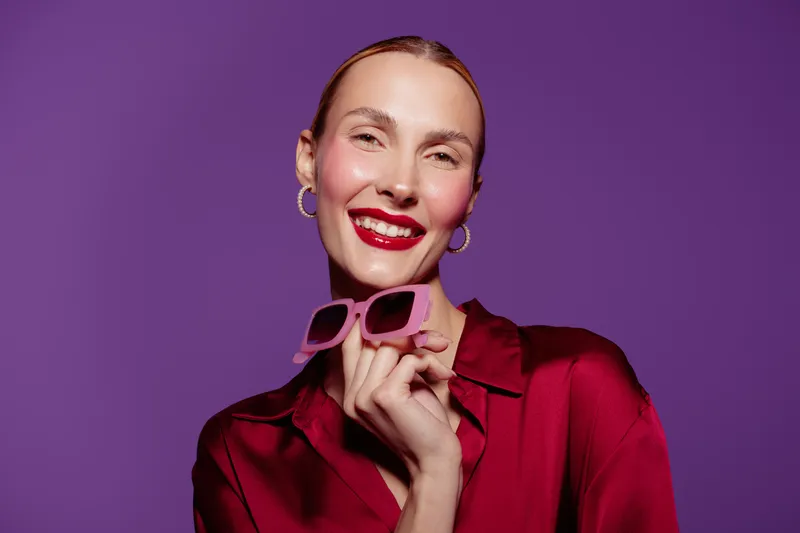 Smiling woman with red lipstick and hoop earrings holds pink sunglasses on purple background. Fashion portrait with bright colors and confident expression in studio lighting.