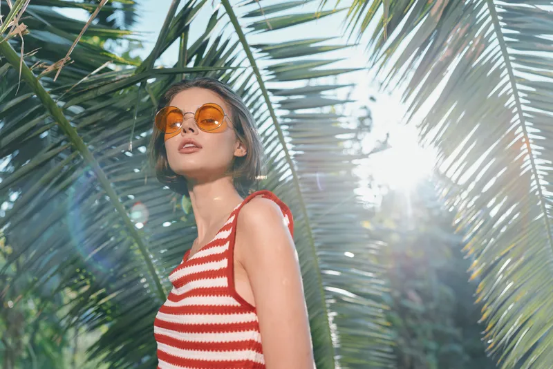 A stylish woman wearing orange sunglasses and a red striped top stands among tropical palm leaves, bathed in sunlight. The vibrant scene conveys a carefree summer mood and bold fashion vibes.