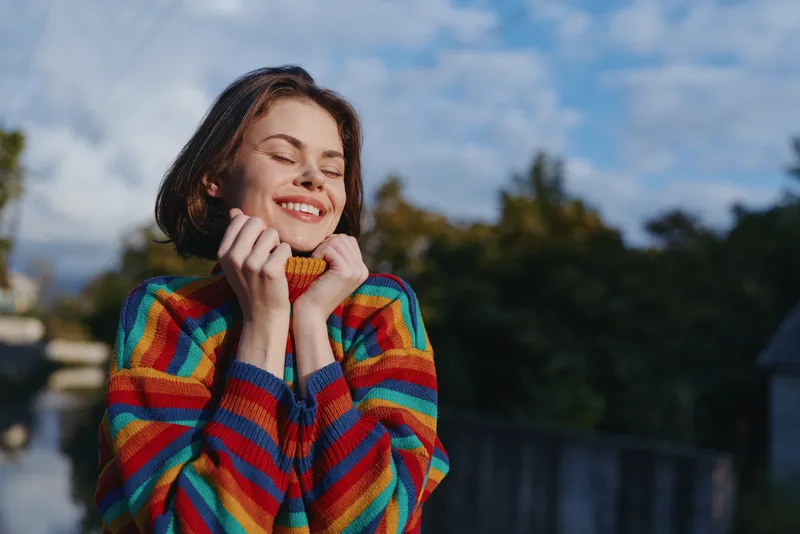 Young woman early 20s with short hair in a colorful striped sweater smiling with eyes closed, hugging collar outdoors by wooden railing in nature. Cozy joyful travel lifestyle.