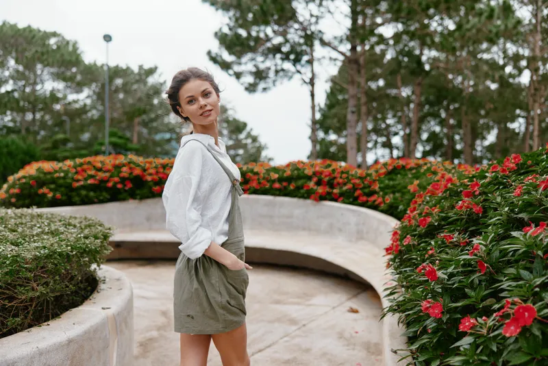 A young woman in overalls stands gracefully in a lush garden filled with vibrant flowers, showcasing a casual summer style and a cheerful expression