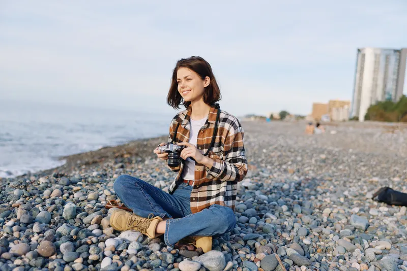 Woman on a pebble beach with a camera, casual plaid shirt and jeans, seated near the sea, capturing moments and enjoying the outdoors under a calm sky