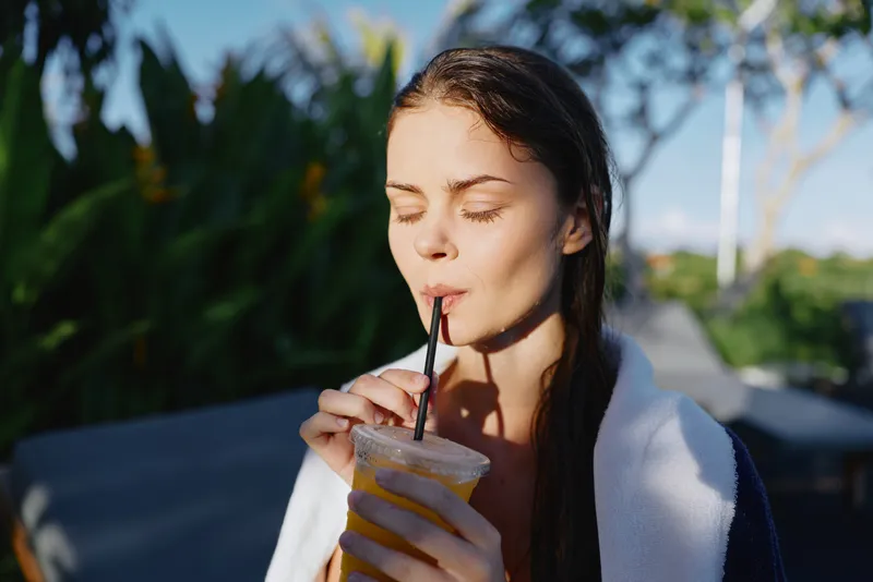 A young woman sips a cold smoothie outdoors, eyes closed, calm and relaxed. Bright sunlight, casual summer lifestyle moment focused on refreshment and self care.