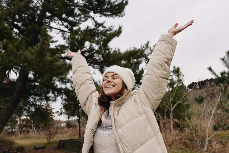 Joyful woman outdoors with arms raised, wearing a beige winter jacket and beanie, enjoying nature and fresh air on a calm, chilly day.