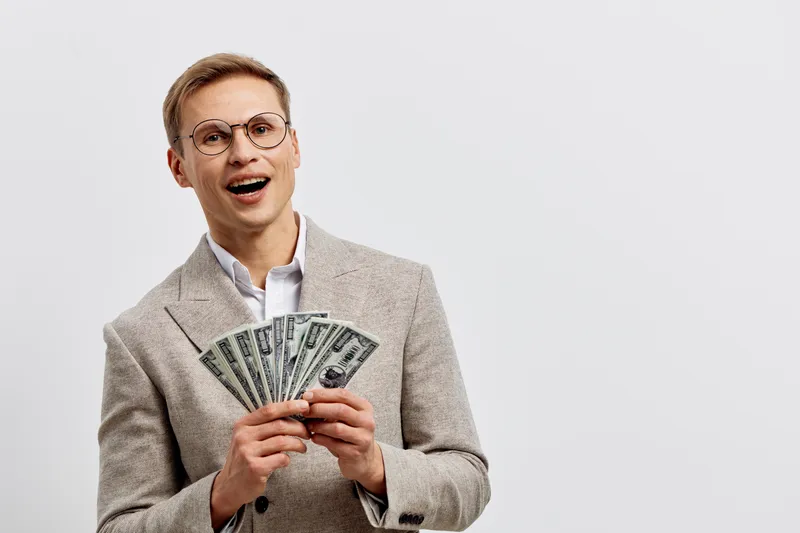 happy young caucasian man hold fan of US dollar cash money wear glasses beige jacket isolated on white background financial success concept