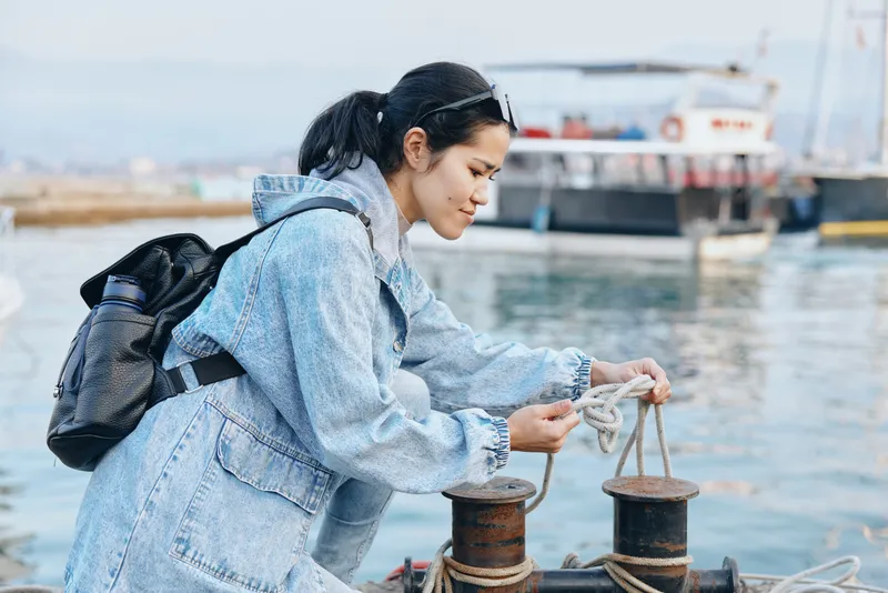 Dock scene with a denim clad figure handling rope at the quay, backpack traveler preparing mooring gear near a calm harbor and boat in the background, morning light on water