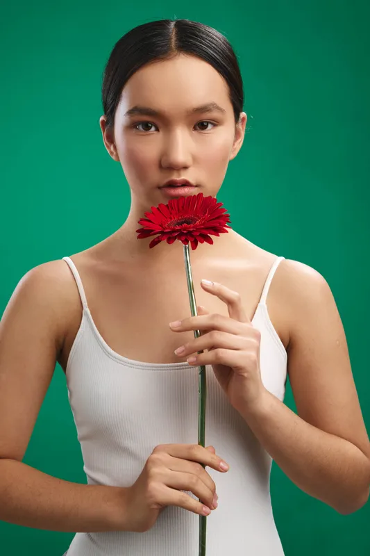 Asian woman stands against a green backdrop, holding a red flower near her lips with a calm gaze, portraying elegance, focus, and subtle emotion.