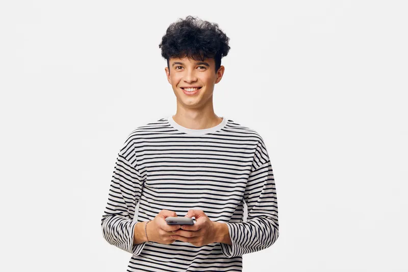 young man in striped shirt smiling while holding smartphone, casual style, curly hair, white background, cheerful expression, modern technology