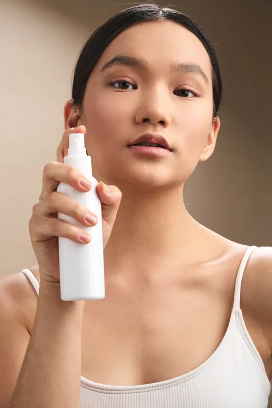 asian woman calmly holds a spray bottle in a bright studio, presenting a skincare routine moment. she smiles softly, exuding calm, clear skin, and a minimalist, modern wellness vibe.