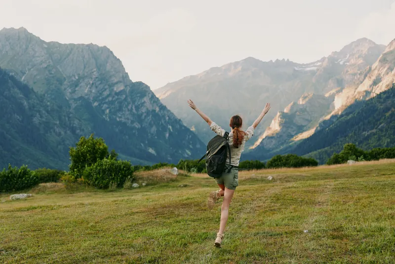 Young woman with backpack running on green meadow in mountains during daytime. Concept of travel, adventure, lifestyle, and freedom in nature with clear sky and bright sunlight.
