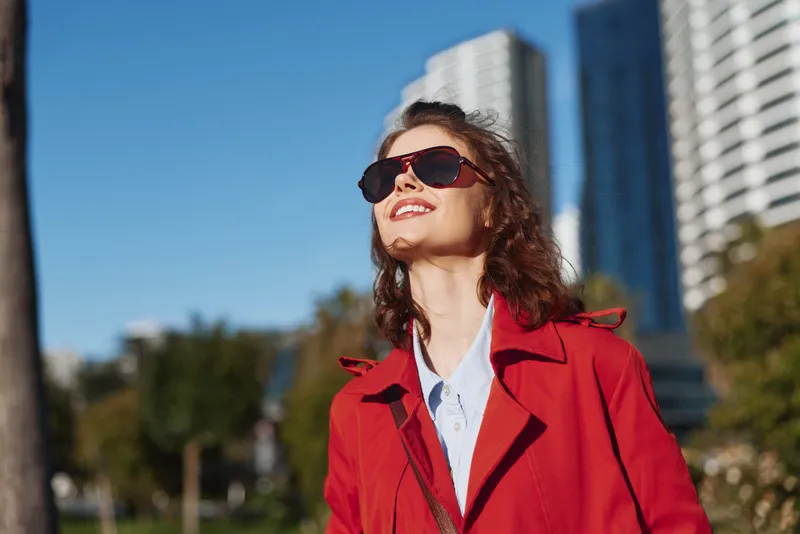Cheerful woman outdoors, wearing a red coat and sunglasses, smiling toward the sun, relaxed posture against a city backdrop, bright daylight and natural colors
