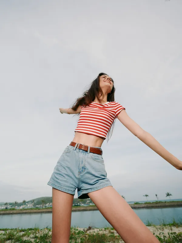 Young woman stretches with open arms in a red striped crop top and denim shorts, outdoors near water, radiating freedom, energy, and carefree summer vibe.