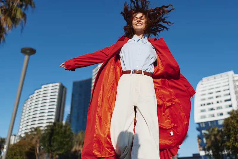Happy woman wearing red coat and white pants jumping outdoors in the city under clear blue sky, expressing joy and freedom on a sunny day.